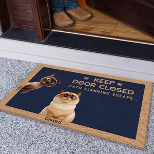 Op de afbeelding: Een blauwe deurmat met de tekst "Keep Door Closed...Cats Planning Escape..." en twee cartoonkatten. De deurmat heeft een bruine jute rand.