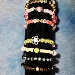 May include: A collection of beaded bracelets in various colors and designs. Some bracelets feature letter beads spelling out words like "PARIS", "POP DEAD", and "DAISY CHAIN". Other bracelets have smiley face and star charms.
