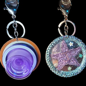 May include: Two keychains with silver clasps. One features a layered design with a purple, white, and brown circular element. The other keychain has a sparkly purple starfish design with a blue beaded border and small white and green flower accents.