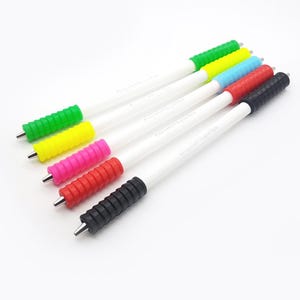 Könnte beinhalten: Sechs weiße Stifte mit verschiedenen farbigen Gummibeschlägen. Die Beschläge sind grün, gelb, rosa, rot, blau und schwarz.