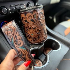 May include: A cylindrical leather cup holder sleeve and a matching leather keychain, both featuring intricate hand-tooled floral scrollwork patterns in brown with black accents and small inlaid turquoise-colored stone details.