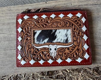 May include: A red, brown, and white leather wallet with a cowhide center panel. The wallet features a carved design with a steer skull and white diamond accents. A zipper and wrist strap are visible.