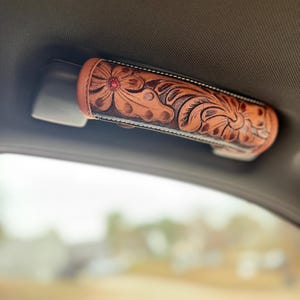 May include: A brown leather grab handle with floral tooling and a red gem center. The handle is attached to a black base and is mounted inside a vehicle. The leather is light brown with dark brown accents.