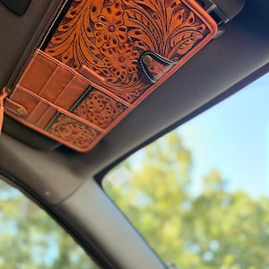 May include: A tan leather car visor organizer featuring intricate floral tooling and small red stone accents. The organizer includes multiple pockets, a zippered compartment, and a snap closure, securely attached to a vehicle sun visor.