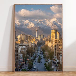 May include: A framed print of a cityscape featuring a wide avenue lined with buildings, leading towards a distant mountain range. The sky is a mix of blue and clouds, with a prominent tower in the center. The buildings are in shades of beige and brown.
