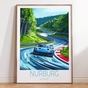 Op de afbeelding: Een ingelijste kunstprint met een blauwe raceauto op een kronkelende baan, omringd door groene bomen en een blauwe lucht. De print bevat het woord "NURBURG" in lichtblauw, met "GERMANY" eronder.