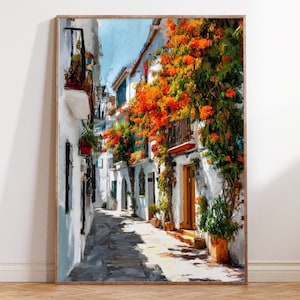 May include: A framed art print showing a narrow street in a European town. White buildings with orange flowers growing down the walls flank the cobbled street. The artwork is vertical and has a wooden frame.