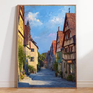 Op de afbeelding: Een ingelijst schilderij toont een Europese straatscène. Het kunstwerk toont kleurrijke gebouwen met rode pannendaken en een geplaveide straat onder een helderblauwe lucht met wollige wolken. Het schilderij heeft een warme, uitnodigende sfeer.