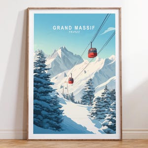 May include: A framed art print showing the Grand Massif in France. The image features red cable cars against a backdrop of snow-covered mountains, with evergreen trees and a clear blue sky. The print is presented in a wooden frame.