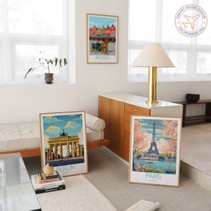 May include: Three framed travel posters with illustrations of cityscapes. The posters are on a white wall in a living room setting. The posters feature illustrations of Bruges, Berlin, and Paris. The posters are all in a similar style with a blue sky and white clouds.