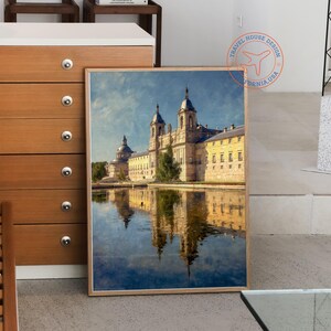 May include: A framed print of a grand building reflected in water. The building has multiple domes and towers, with a yellow facade and a blue sky background. The print is in a light brown frame.