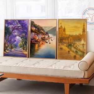 May include: Three framed watercolor prints. The left print depicts a street lined with purple flowering trees. The middle print shows a coastal town. The right print features a European building with a fountain.