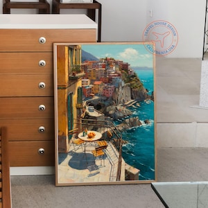 Op de afbeelding: Een ingelijste prent van een Italiaans kustdorpssc&egrave;ne. Het kunstwerk toont kleurrijke gebouwen op een klif met uitzicht op de turquoise zee. Een kleine tafel en stoelen staan op een balkon op de voorgrond, met sinaasappels op de tafel.