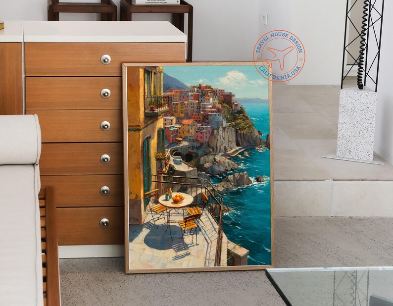 Op de afbeelding: Een ingelijste prent van een Italiaans kustdorpssc&egrave;ne. Het kunstwerk toont kleurrijke gebouwen op een klif met uitzicht op de turquoise zee. Een kleine tafel en stoelen staan op een balkon op de voorgrond, met sinaasappels op de tafel.