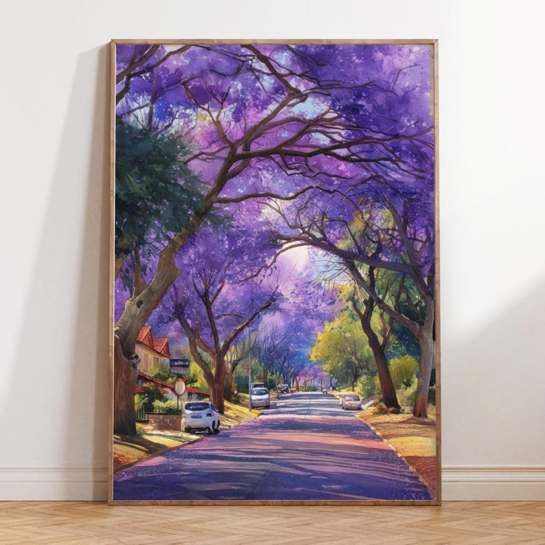 May include: A framed watercolor painting of a tree-lined street with purple flowering trees. The artwork depicts a sunny day with shadows cast on the road. Cars are parked along the street, and houses are visible in the background. The painting is in a wooden frame.