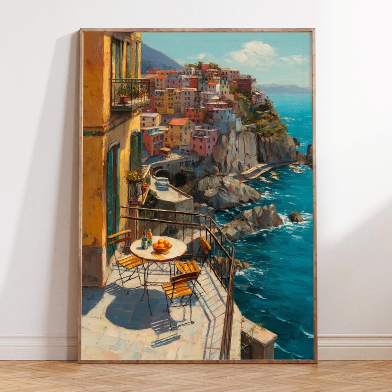 Op de afbeelding: Een ingelijste prent van een Italiaans kustdorp. Het kunstwerk toont kleurrijke gebouwen op een klif met uitzicht op de turquoise zee. Een balkon met een tafel en stoelen staat op de voorgrond. De prent zit in een houten lijst.
