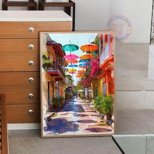 May include: A framed watercolor painting depicts a vibrant street scene. Colorful umbrellas float above a narrow street lined with buildings in shades of yellow, orange, and pink. The artwork evokes a sunny, tropical atmosphere.