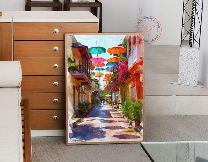 May include: A framed watercolor painting depicts a vibrant street scene. Colorful umbrellas float above a narrow street lined with buildings in shades of yellow, orange, and pink. The artwork evokes a sunny, tropical atmosphere.