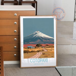 May include: A framed art print featuring a landscape scene of Cotopaxi National Park. The artwork depicts a snow-capped mountain under a blue sky, with a dirt road leading to small buildings. The text "Cotopaxi National Park" is at the bottom.