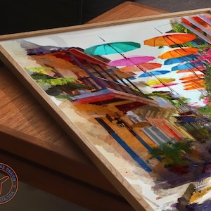 May include: A framed watercolor painting depicts a vibrant street scene with colorful umbrellas suspended overhead. The artwork showcases buildings with orange, blue, and red hues, complemented by lush greenery and bright pink flowers. The frame is made of light-colored wood.