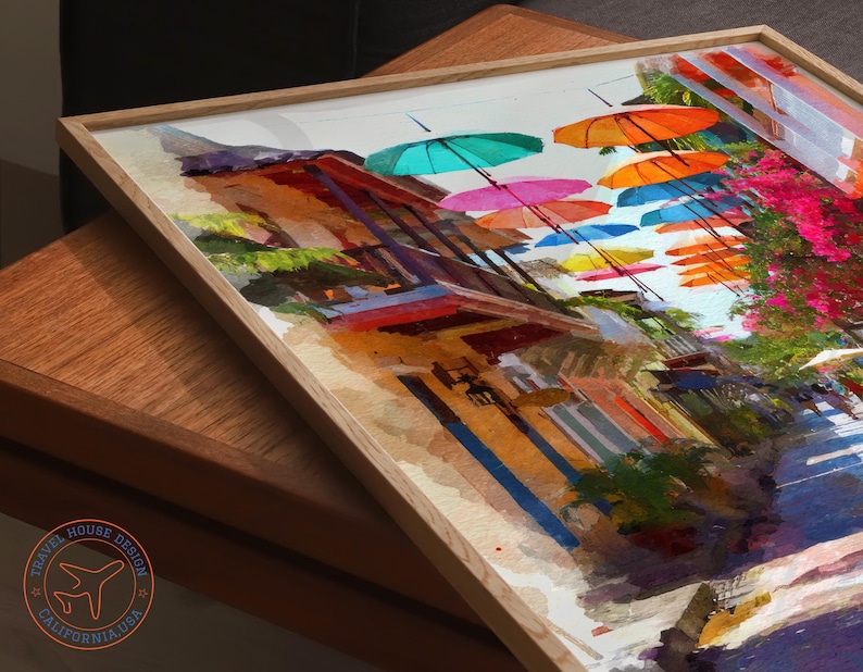 May include: A framed watercolor painting depicts a vibrant street scene with colorful umbrellas suspended overhead. The artwork showcases buildings with orange, blue, and red hues, complemented by lush greenery and bright pink flowers. The frame is made of light-colored wood.
