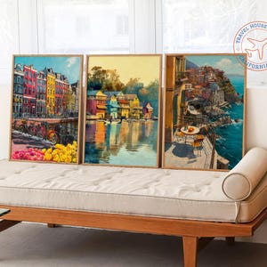 Op de afbeelding: Drie ingelijste kunstdrukken met Europese stadsgezichten. Het kunstwerk toont kleurrijke gebouwen, waterreflecties en kustgezichten. De prints worden tentoongesteld op een houten daybed. Het Travel House Design-logo staat rechtsboven.