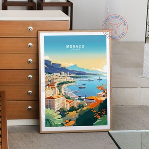 May include: A framed art print featuring a colorful illustration of Monaco, with the text "MONACO EUROPE" at the top. The artwork depicts a coastal city with buildings, boats, and mountains under a blue sky and a setting sun. The print is in a wooden frame.