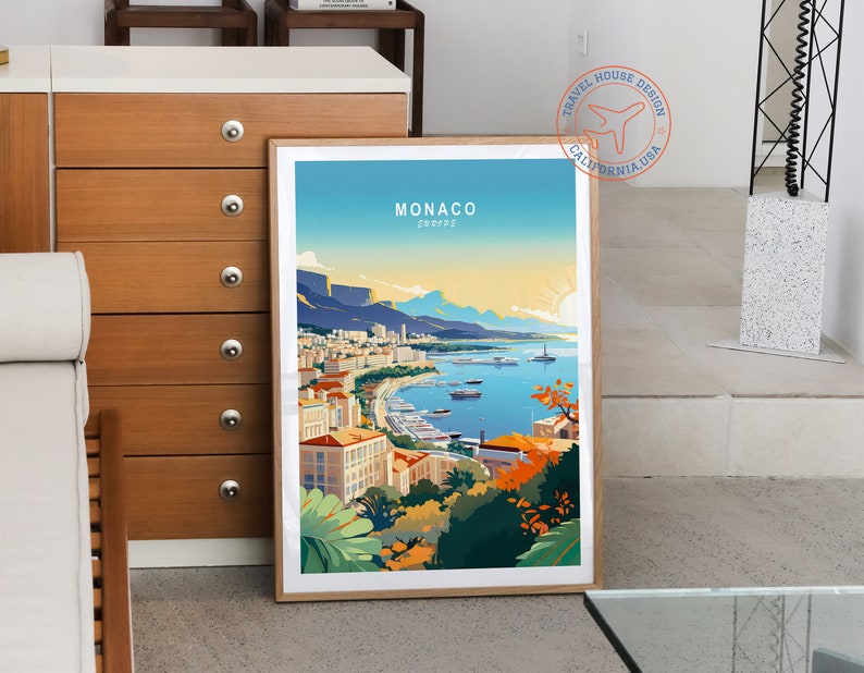 May include: A framed art print featuring a colorful illustration of Monaco, with the text "MONACO EUROPE" at the top. The artwork depicts a coastal city with buildings, boats, and mountains under a blue sky and a setting sun. The print is in a wooden frame.