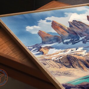 K&ouml;nnte beinhalten: Aquarell-Kunstdruck eines Bergpanoramas mit schneebedeckten Gipfeln, einem t&uuml;rkisfarbenen See und einem blauen Himmel mit Wolken. Das Kunstwerk ist in einem hellen Holzrahmen gerahmt. Das Logo von Travel House Design ist sichtbar.