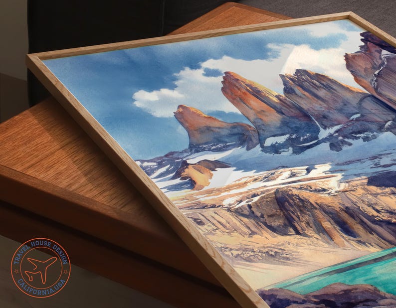 K&ouml;nnte beinhalten: Aquarell-Kunstdruck eines Bergpanoramas mit schneebedeckten Gipfeln, einem t&uuml;rkisfarbenen See und einem blauen Himmel mit Wolken. Das Kunstwerk ist in einem hellen Holzrahmen gerahmt. Das Logo von Travel House Design ist sichtbar.