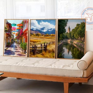 May include: Three framed landscape art prints. One depicts a colorful street with umbrellas, another a field with mountains, and the third a canal with a tower. The frames are light wood, and the art is displayed on a wooden daybed.