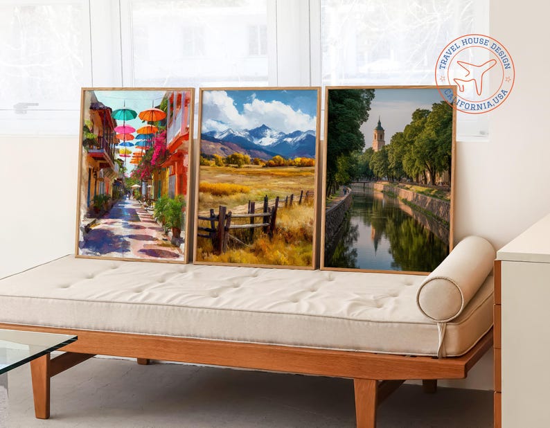 May include: Three framed landscape art prints. One depicts a colorful street with umbrellas, another a field with mountains, and the third a canal with a tower. The frames are light wood, and the art is displayed on a wooden daybed.