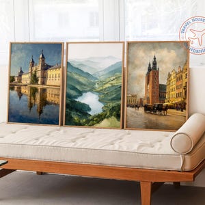 May include: Three framed watercolor prints featuring European landscapes. The prints depict a monastery reflected in water, a mountain valley with a river, and a city scene with a tall building and horse-drawn carriages. The frames are made of light-colored wood.