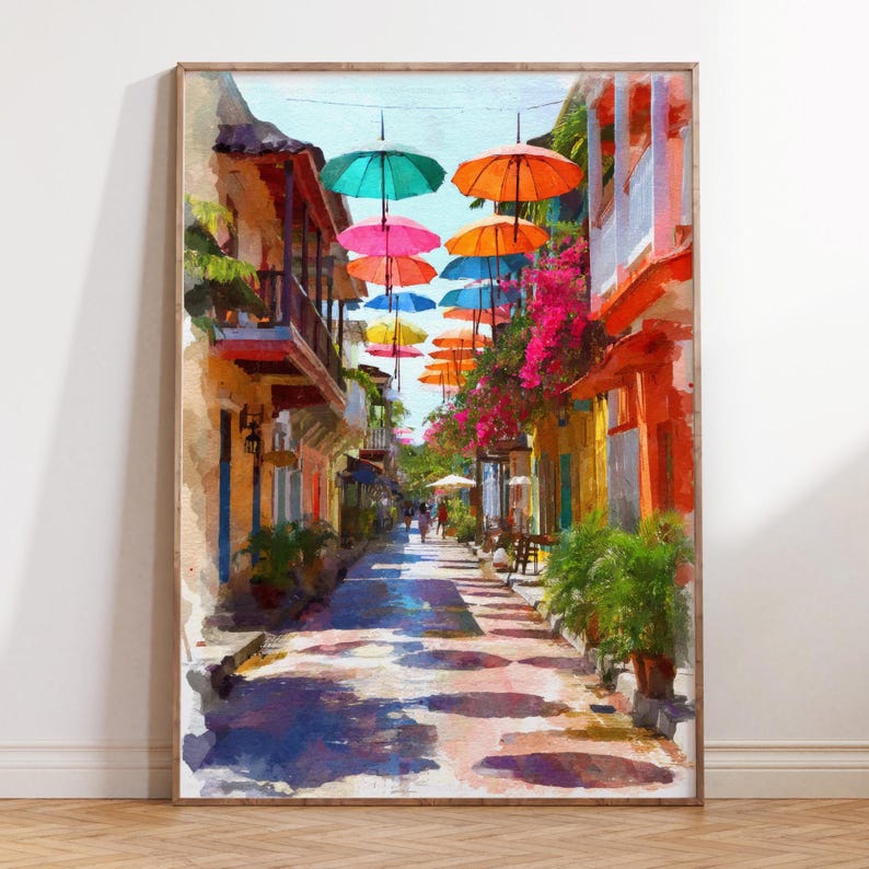 May include: A framed watercolor painting of a colorful street scene. The artwork depicts a narrow street lined with buildings in shades of orange, yellow, and pink. Overhead, vibrant umbrellas in various colors are suspended, casting shadows on the cobblestone street.
