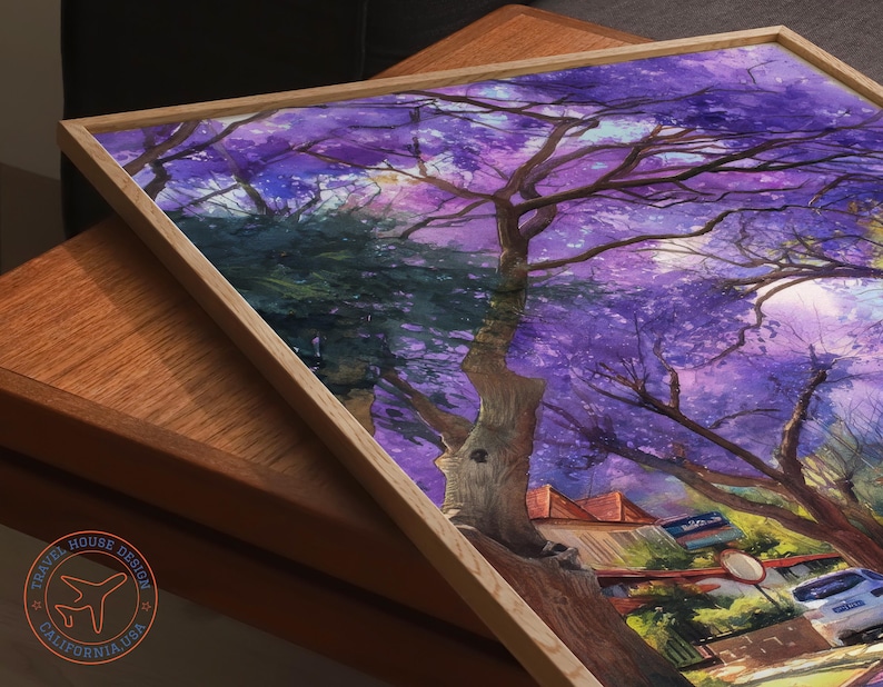 May include: A framed art print featuring a vibrant watercolor painting of a tree with purple blossoms. The artwork is set against a backdrop of buildings and a street scene, with a light-colored wooden frame. The Travel House Design logo is visible.