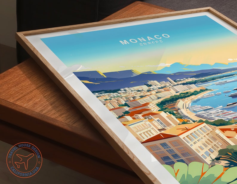 May include: Framed art print of Monaco, Europe, featuring a vibrant illustration of the city's coastline, buildings, and harbor. The print has a light wood frame and white matting. The artwork is signed by Travel House Design, California, USA.