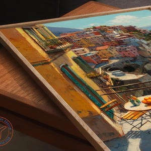Op de afbeelding: Een ingelijst schilderij van een Italiaans kustdorp met kleurrijke gebouwen, een blauwe zee en een tafel met sinaasappels en drankjes. Het kunstwerk zit in een licht houten lijst en rust op een houten oppervlak.