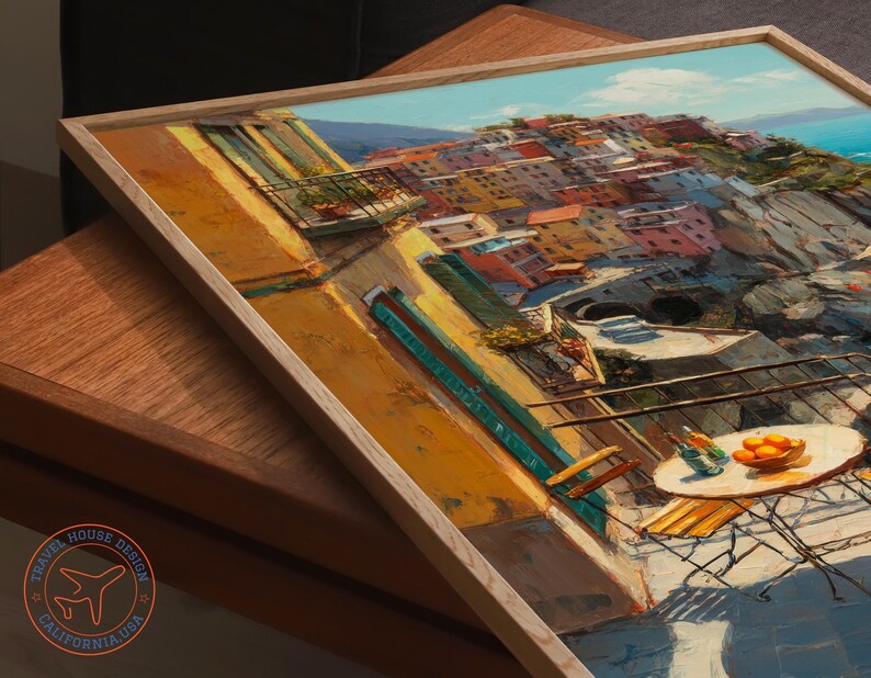 Op de afbeelding: Een ingelijst schilderij van een Italiaans kustdorp met kleurrijke gebouwen, een blauwe zee en een tafel met sinaasappels en drankjes. Het kunstwerk zit in een licht houten lijst en rust op een houten oppervlak.