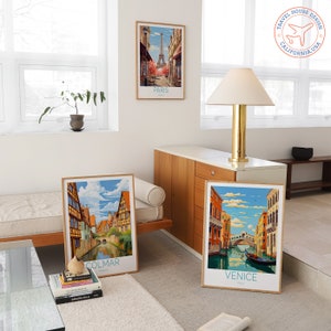 Puede incluir: Tres p&oacute;sters de viaje vintage con ilustraciones de Par&iacute;s, Colmar y Venecia. Los p&oacute;sters est&aacute;n enmarcados en madera y tienen un fondo blanco con texto azul y blanco. Los p&oacute;sters se muestran en una pared blanca en una sala de estar moderna.