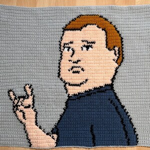 May include: Crocheted portrait of a cartoon character with brown hair, a blue shirt, and a pink hand making a rock and roll hand gesture. The background is gray.