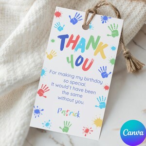 May include: A white gift tag with the words "THANK YOU" in colourful letters, surrounded by handprints and paint splatters. The tag includes a message and the name "Patrick". A natural twine string is attached.