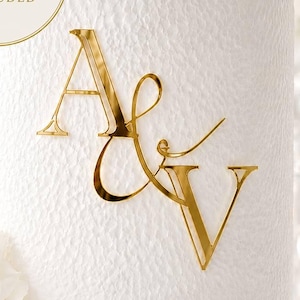 Puede incluir: Una tarta blanca con una superficie texturizada, adornada con las iniciales doradas "A & V". La tarta está sobre un soporte blanco, con flores blancas y una vela en el fondo. También es visible un círculo dorado con el texto "FREE CAKE CHARM HOLDER INCLUDED".