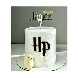 May include: A white cake with a gold "James" cake topper and a lightning bolt. The cake has a black "Hp" design and a small owl figurine. The cake sits on a white cake board.