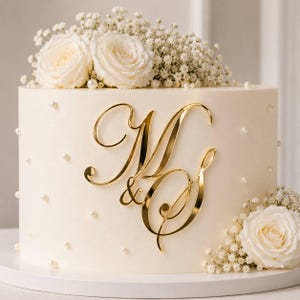 Könnte beinhalten: Eine weiße Hochzeitstorte, verziert mit goldfarbenen Initialen "M & G" und kleinen Perlenakzenten. Die Torte ist mit weißen Rosen und Schleierkraut geschmückt. Die Torte steht auf einem weißen Kuchenständer.