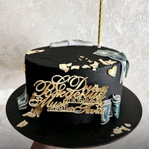 May include: A black birthday cake decorated with gold leaf, faux money, and a gold candle. The cake has a gold script message on the front. The cake sits on a black cake board.