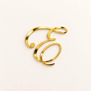 May include: A gold-colored letter E in a cursive script font. The letter features a reflective, metallic finish with smooth, flowing lines that create a decorative shape. The design is characterized by its elegant, interconnected loops and curves.