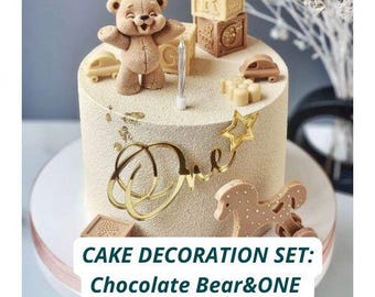 May include: A beige cake decorated with a brown teddy bear, toy blocks, and a gold star. The cake has a gold "One" cake topper and a candle. The text on the cake reads "CAKE DECORATION SET: Chocolate Bear&ONE."