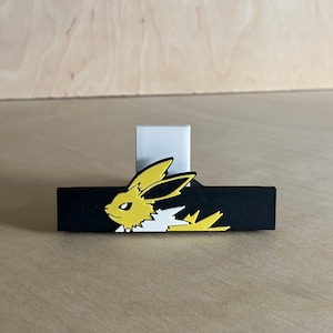 May include: A black and white phone stand with a yellow and white cartoon character on the front. The character is a Pikachu from the Pokemon franchise.