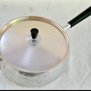 May include: A vintage aluminum saucepan with a lid and a black handle. The pan has a round shape with a lid and a small black knob. A gold-colored ring is attached to the side. The handle is black and angled upwards.