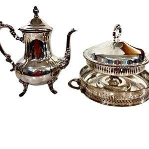 May include: A silver-colored tea set featuring a teapot with a curved handle and a covered serving dish. The teapot has a decorative spout and sits on three small feet. The serving dish has a decorative rim and a handle on top.
